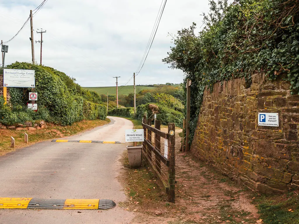 A road on the left with black and yellow speed bumps, and the path on the right next to a stone wall.