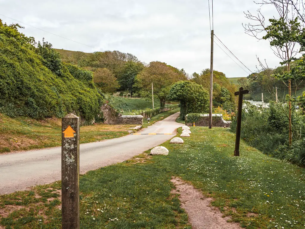 A road on the left and green path of grass to the right and a wooden stump trail signpost with a yellow arrow pointing ahead.