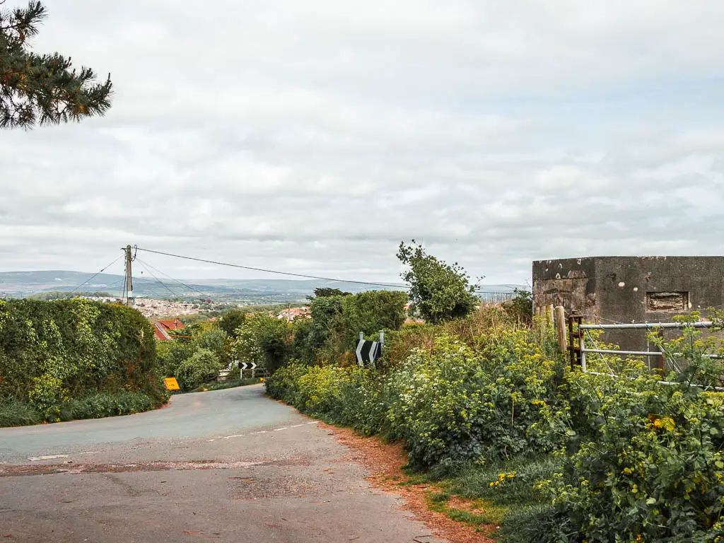 a road going straight and down, with some bushes on the right and hedges on the left and a view to a village in the distance. 