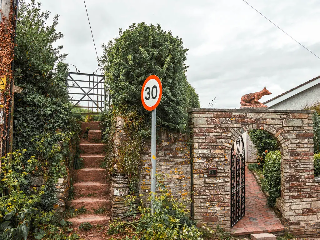 Uphill stone steps on the left and a stone archway entrance to a home on the right. There is a fox stature on top of the arch.