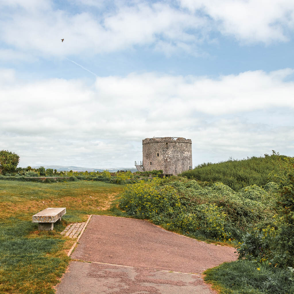 A bench on the green on the left, bushes on the right and Mount Batten ahead. 