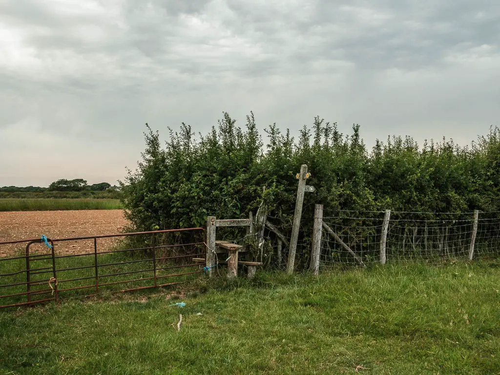 A metal gate and wooden style, with a trail signpost and hedge on the right.