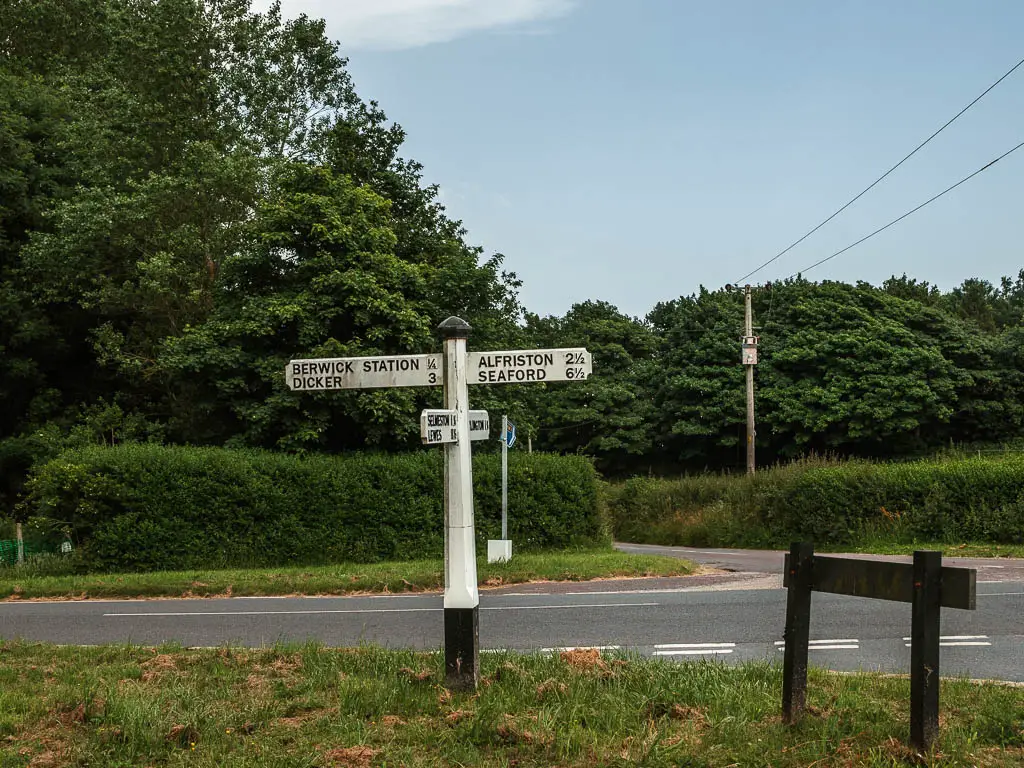 A white sign post on the side of the road, pointing to Alfriston to the right and Berwick Station to the left, at the end of the Long Man of Wilmington walk.