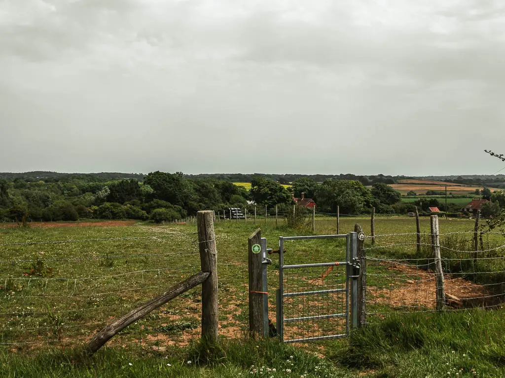 A barbed wire fence and metal gate leading to a field. There are trees on the other side of the field. 