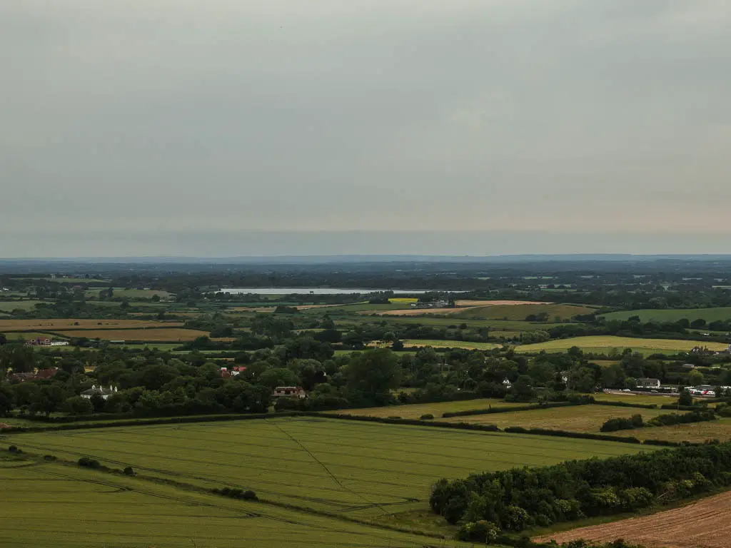 A view across the flat landscape of many fields , separated by trees and the Arlington reservoir in the distance, on the walk to the Long Man of Wilmington