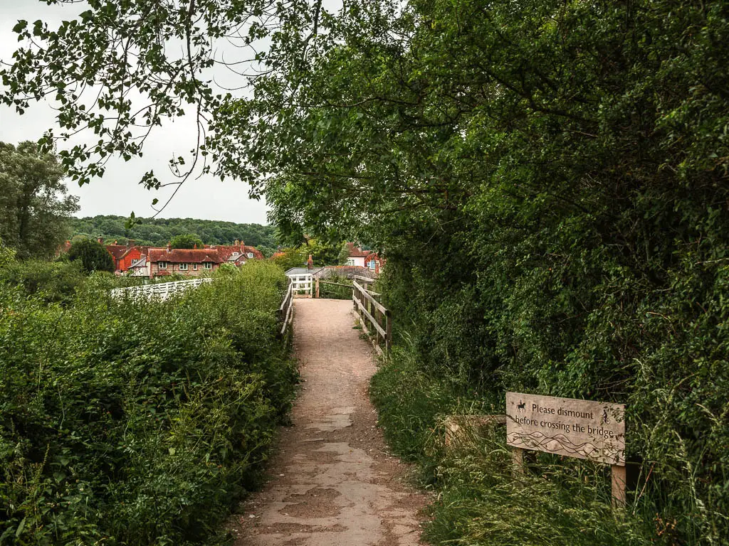 A path leading to a brush, lined with bushes and trees, and a wooden sign on the right, on the walk into Alfriston.