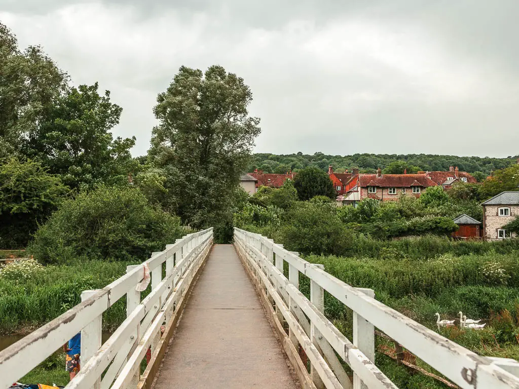 A bridge with white wooden trailings leading to more greenery and the rooftops of Alfriston ahead to the right.