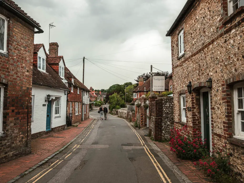 A residential road in Alfriston, lined with shoes made of brick and flint. There are a couple of people walking ahead on the road. 