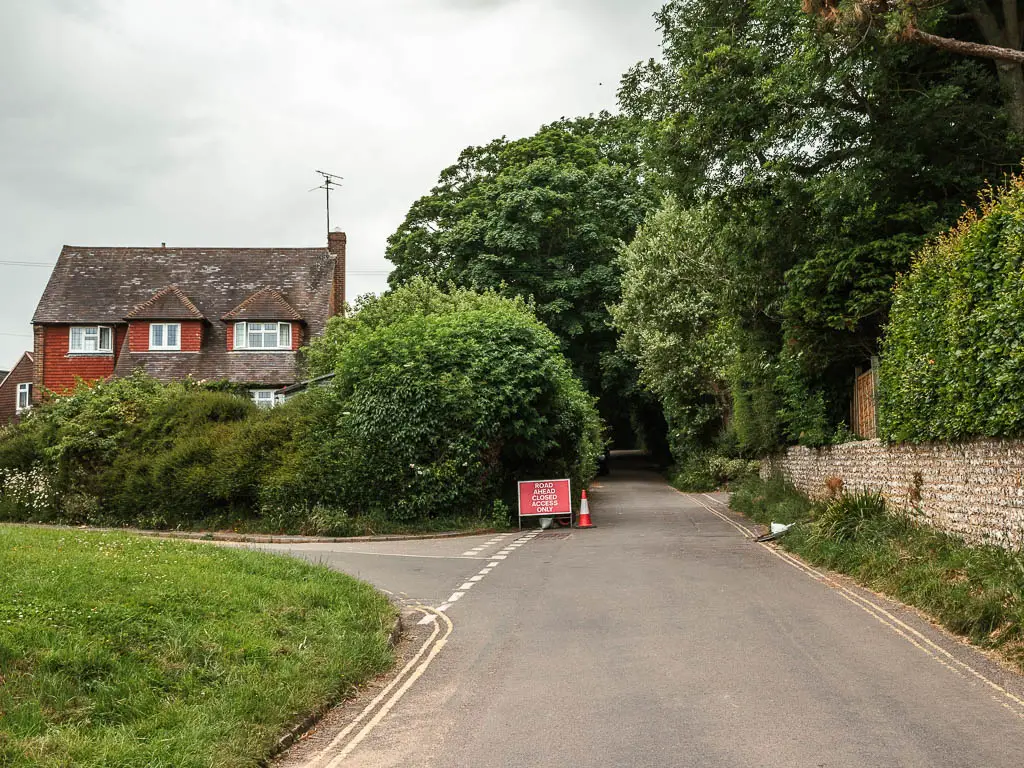 The road as it runs straight, with a green and side road to the left and a house partially hidden behind the hedge on the left. There is a stone wall on the right with hedges on top.