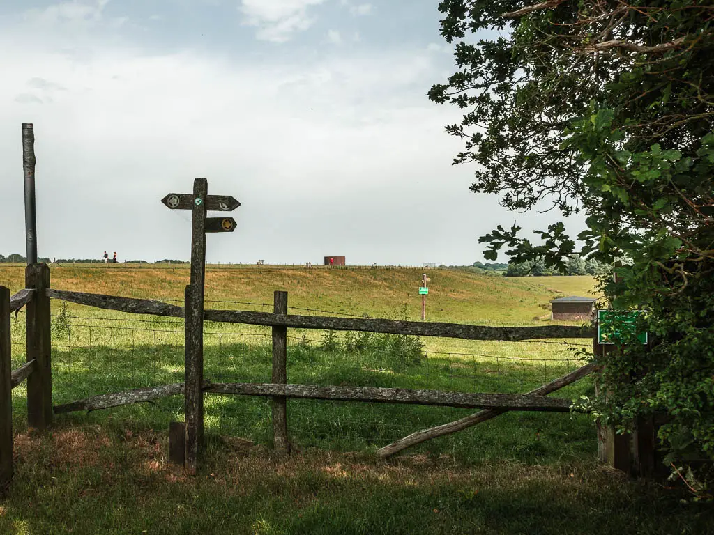 A wooden fence with wires, and a wooden trail signpost, and a field on the other side. 