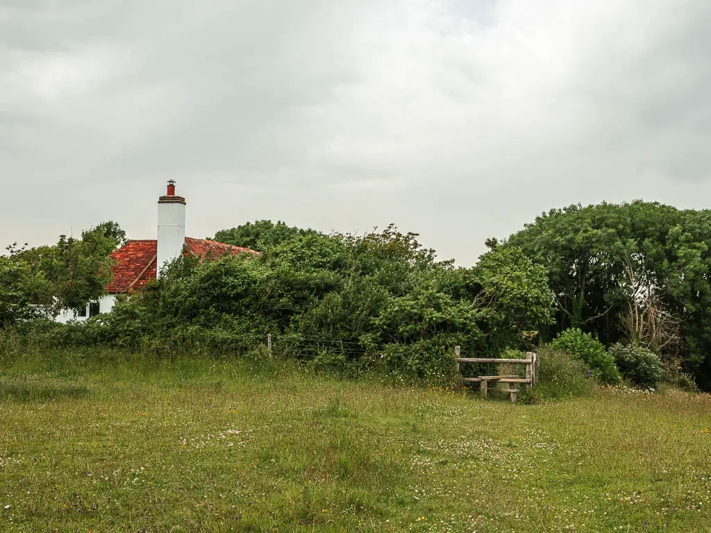 A field, with bushes and a red rooftop on the other side of them, and a wooden stile just to the right.