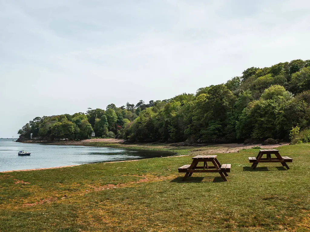 Looking across the green to the sea and woodland trees ahead. There are a couple of wooden picnic benches on the green.