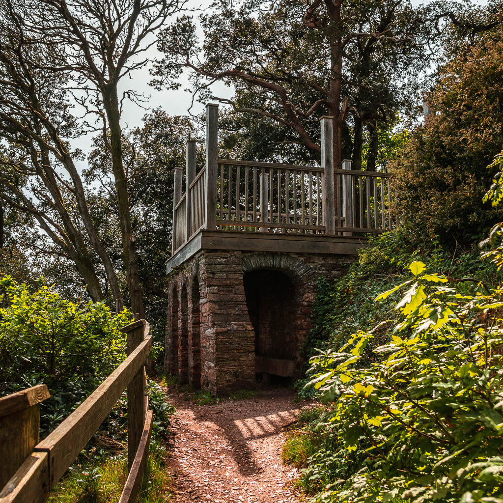 A stone structure on the path, with a wooden fence on the left and some bushes on the right.