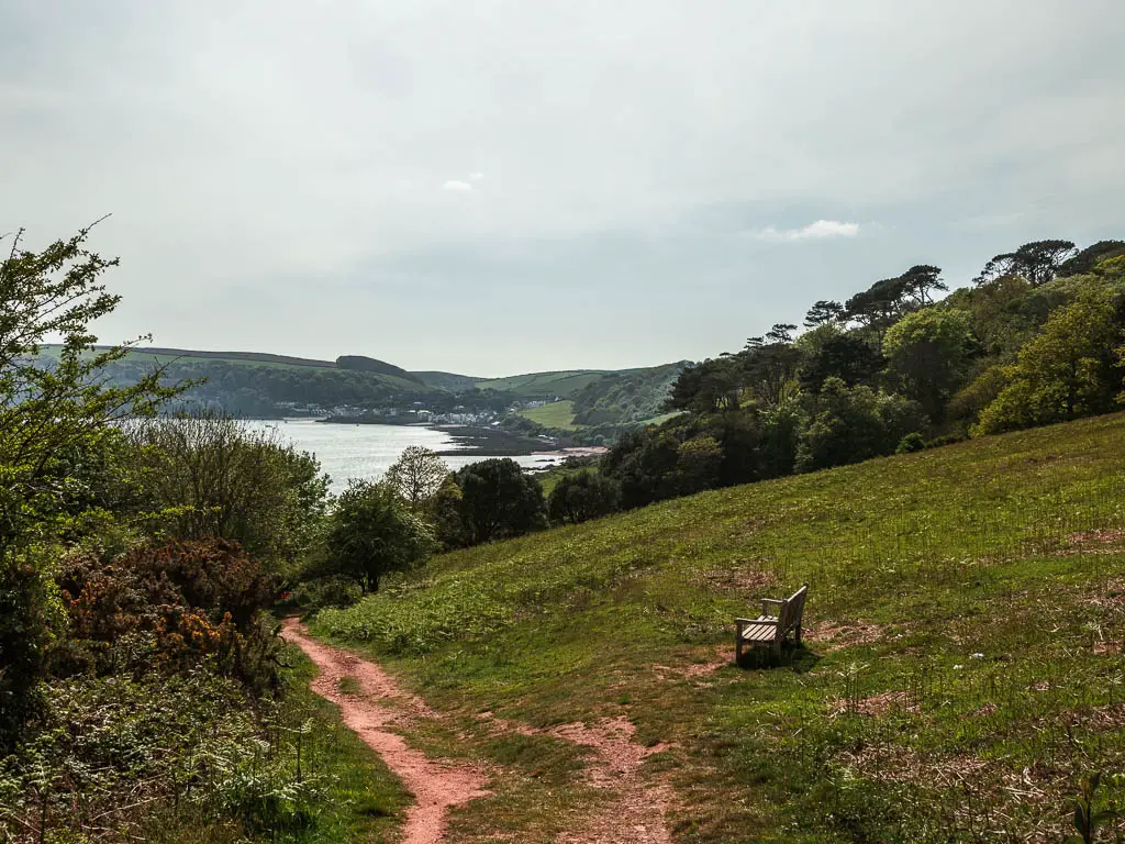 A dirt trail leading down a grass hill with a view to the sea and Kingsand in the distance, on the circular walk through Mount Edgcumbe. There is a wooden bench on the grass hillside. 