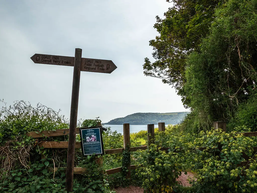 A wooden trail signpost in front of a wooden gate and bushes, with a view to the sea in the distance on the other side. 