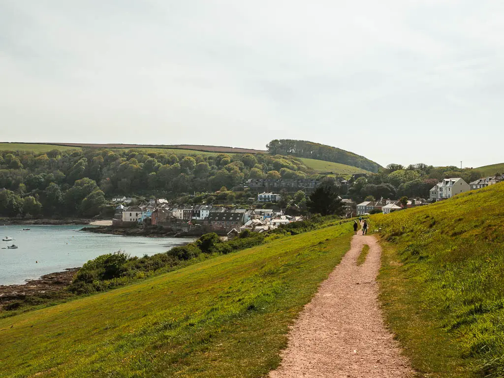 A dirt path along the side of a hill, leading to the village of Kingsand by the sea ahead, on the circular walk through Mount Edgcumbe country park. There are two people walking ahead on the path. 