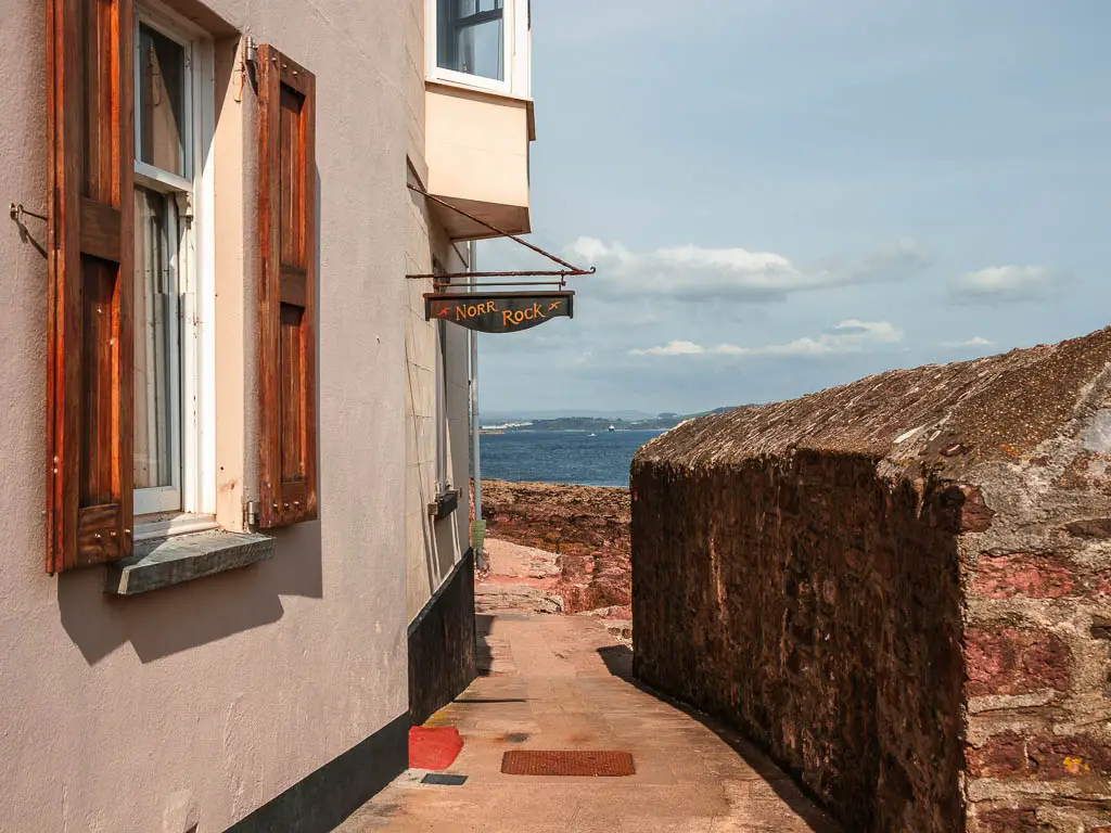 Looking along a small path, with a stone wall to the right and building to the right on the circular walk through Kingsand and Mount Edgcumbe country park. The sea is visible in the distance ahead. 