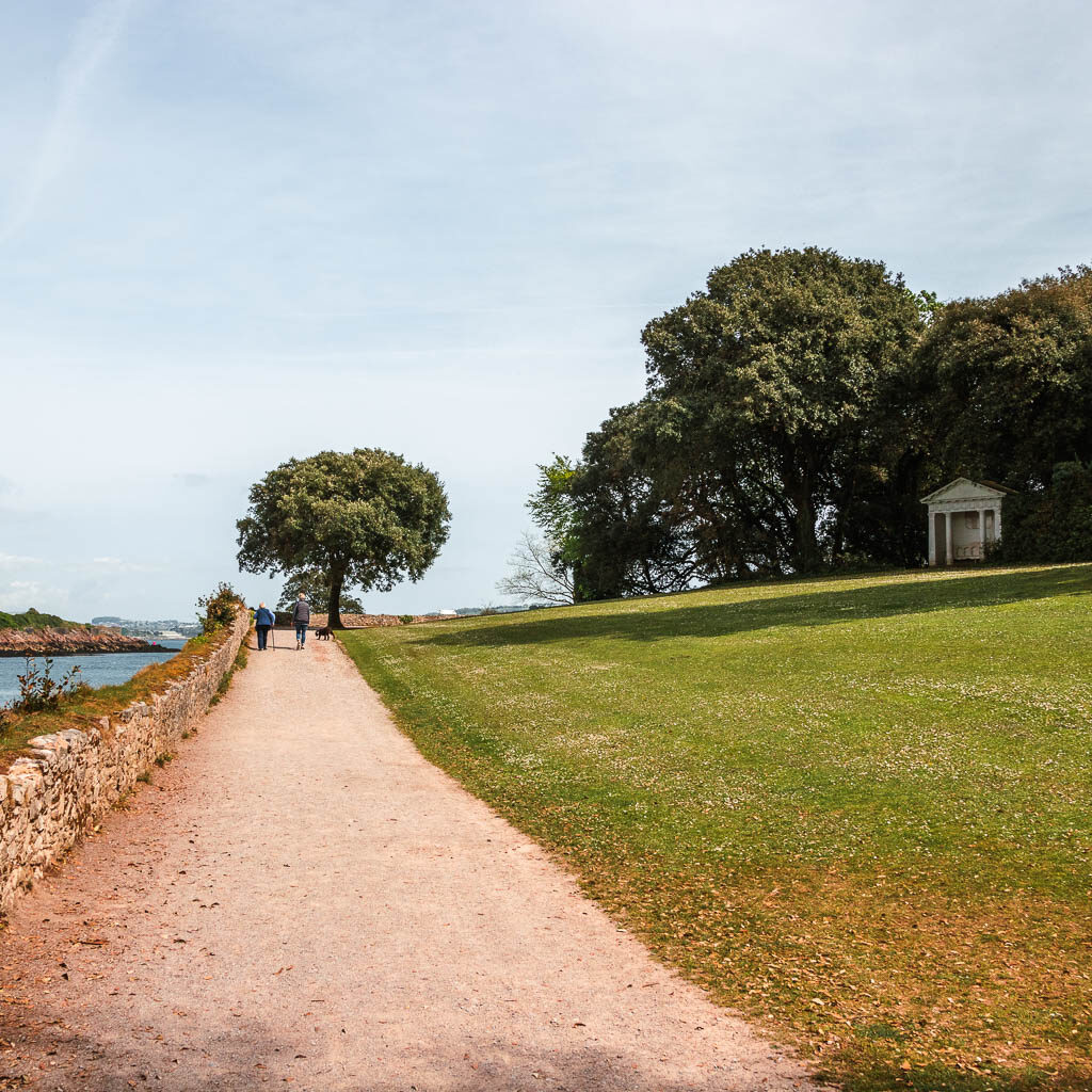 A wide path on the left and neatly cut green on the right. There is a stone wall to the left of the path and some people walking ahead in the distance. 