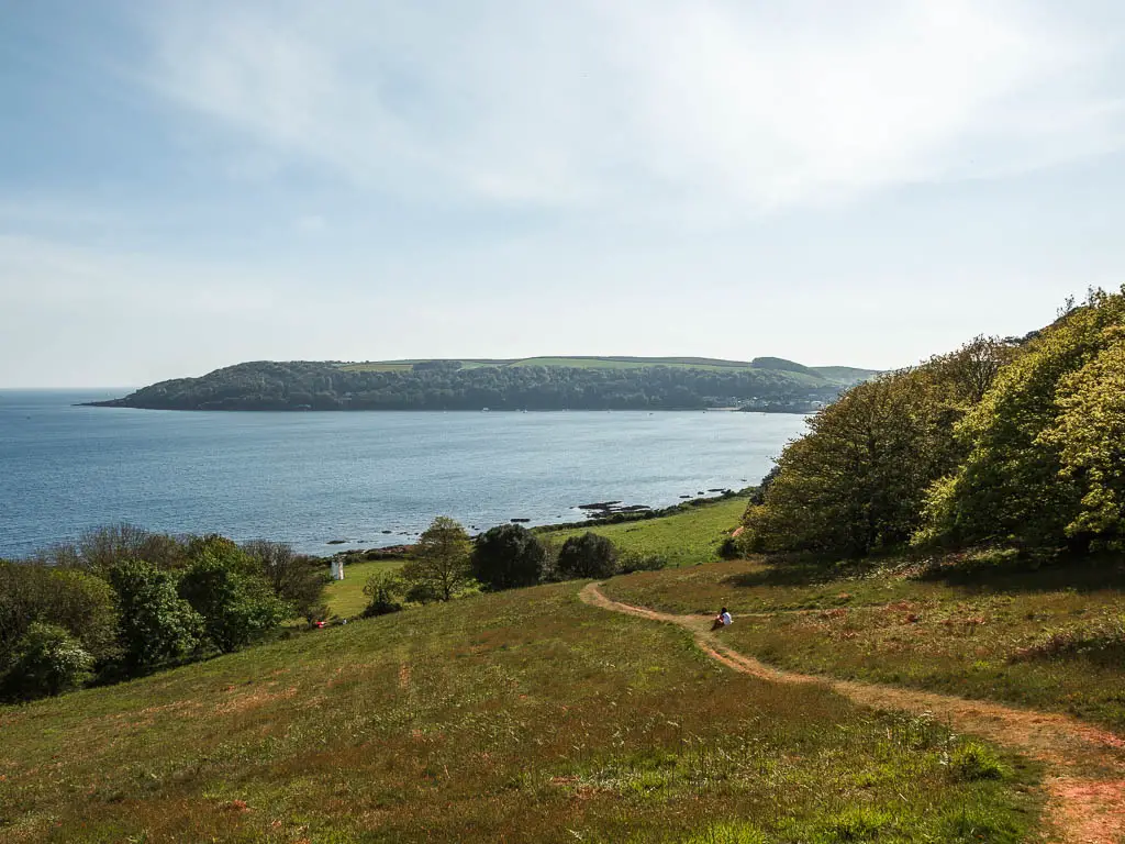 Looking down the grass hill to the blue sea below. There is a trail running down the hill, and a person sitting next to it.