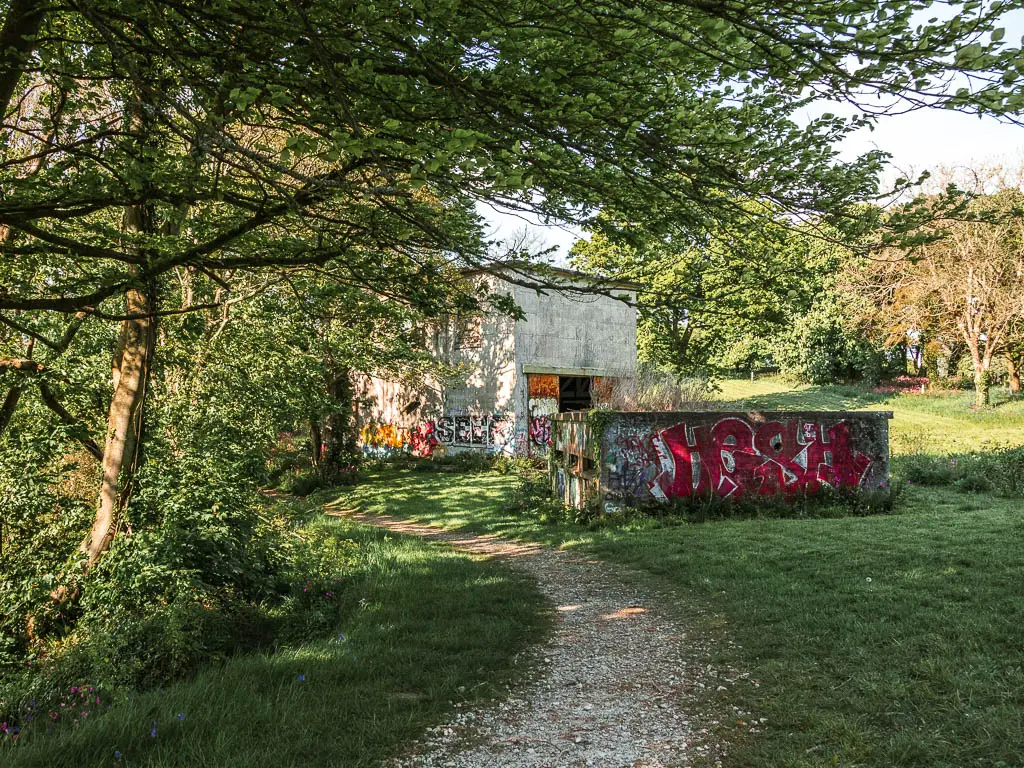 A gravel path through the green grass with a couple of abandoned buildings covered in graffiti ahead. 