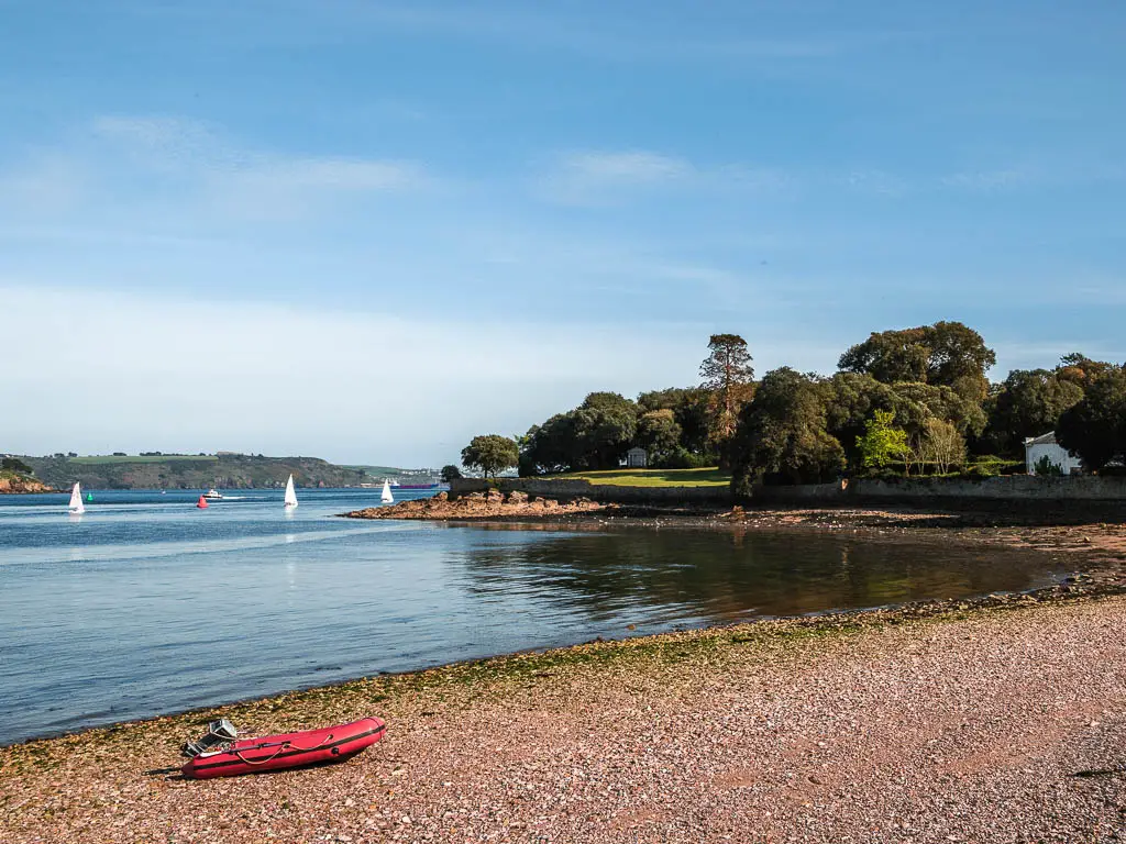 Looking across the shingle beach to the clam water at the start of the Mount Edgcumbe to Kingsand circular walk. There is a small red dingy on the beach.