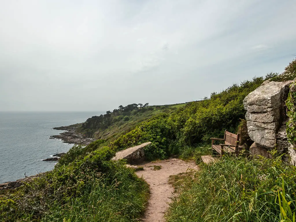 A trail at the top of a hill on the walk from Mousehole to Lamorna. There is a wooden bench on the right, next to a rock, and view down to the sea and coastline to the left.