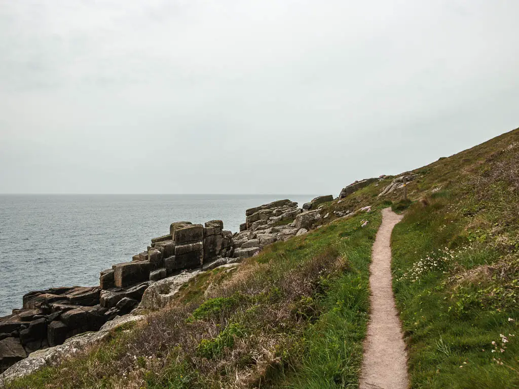 A narrow trail along the side of a grass covered hill on the coastal walk from Mousehole to Lamorna. There are large rocks at the bottom of the hill on the left next to the sea.