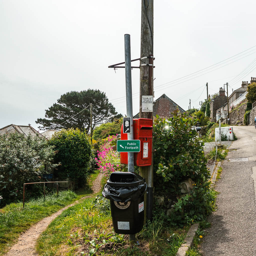 A road leading uphill to the right, and a trail to the left. There is a black bin and red post box in between them.