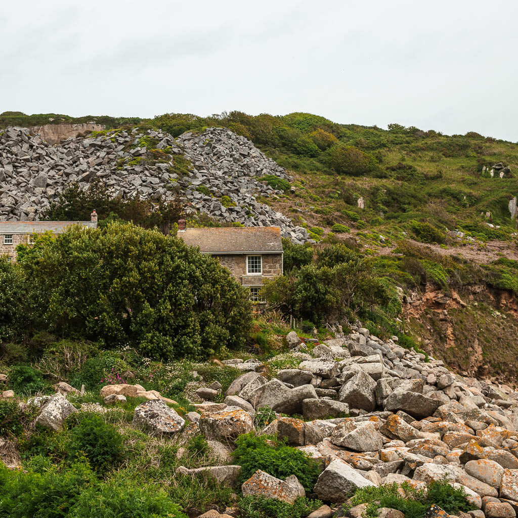 A pile of rocks leading towards a small cottage partially hidden behind the bush, half way through the walk from Mousehole to Lamorna.