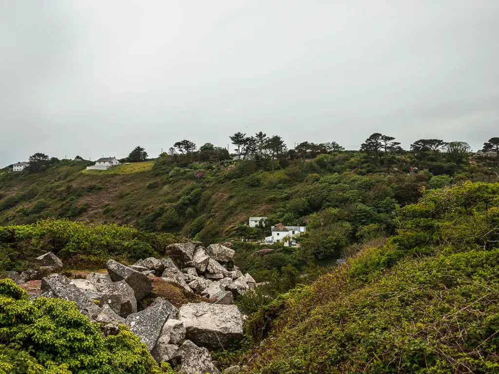 Looking across the bushes and rocks to the grass and bushes covered hill on the other side. There is a white home nestled between some bushes on the hill.