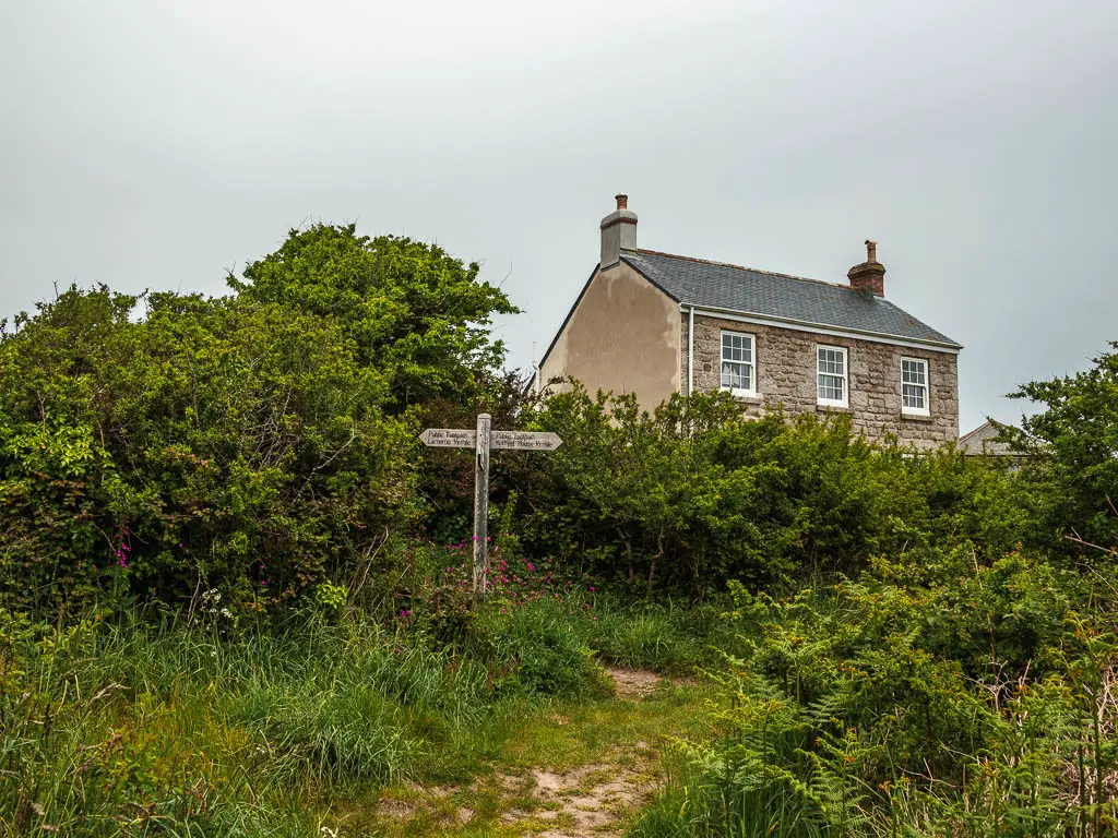 A wooden trail signpost infant of bushes, with a house to the right. The grass in front of the sign is tall and wild.