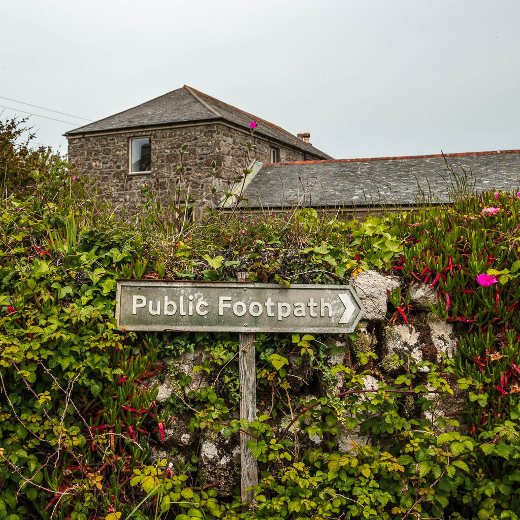 A public footpath sign on the walk from Mousehole to Lamorna. The sign is in front of a stone wall covered in ivy. There is a stone house behind the wall.