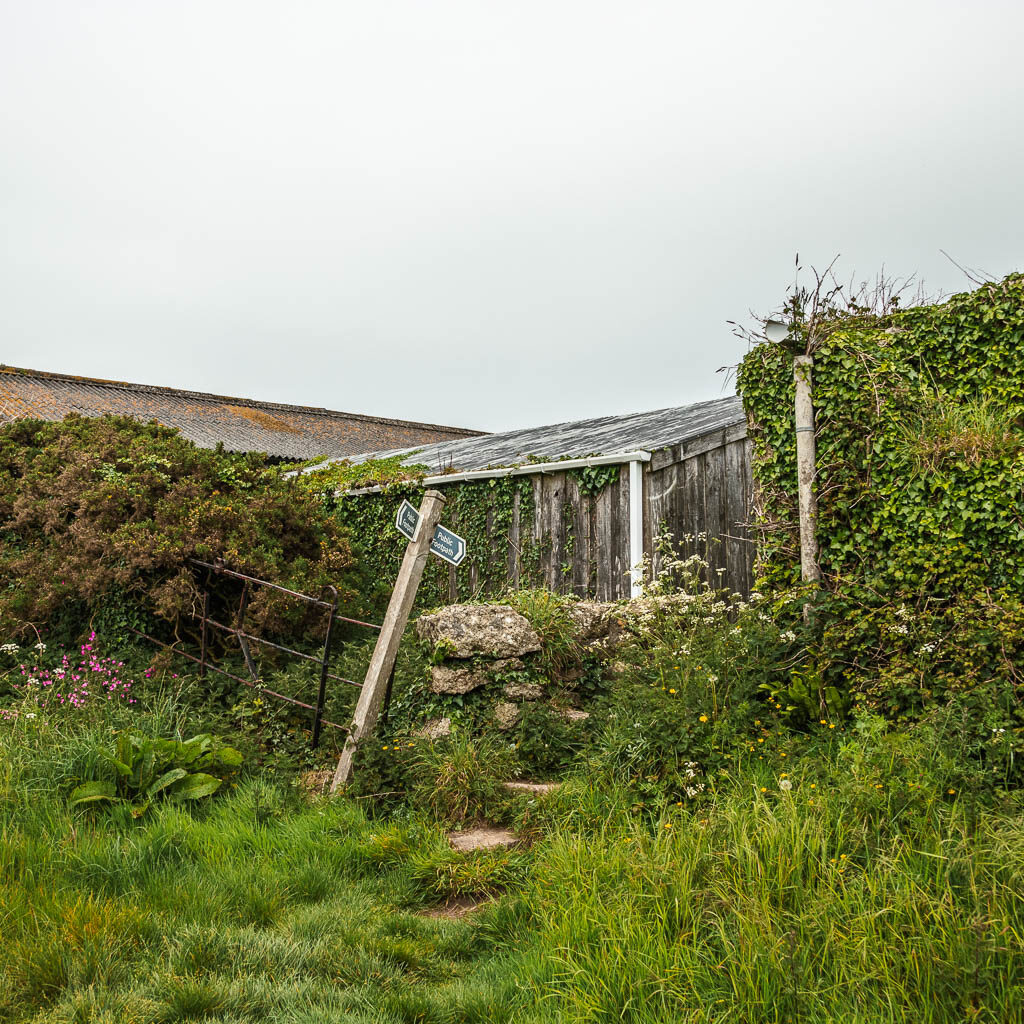 Tall grass in front of a stone wall and trail signpost, pointing in between the farm buildings.