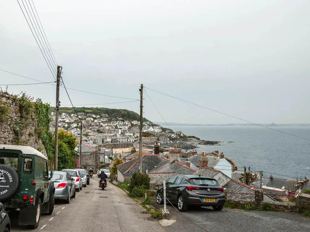 The road leading down towards the village of Mousehole at the end of the walk back from Lamorna. There are cars parked on the left of the road. There is a man on a motorbike driving down the road. 