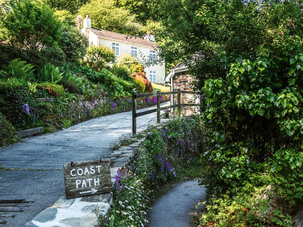 A road leading uphill on the left and a small path on the right. There is a coast path sign pointing to the right trail. The trail is lined with bushes on the right side.