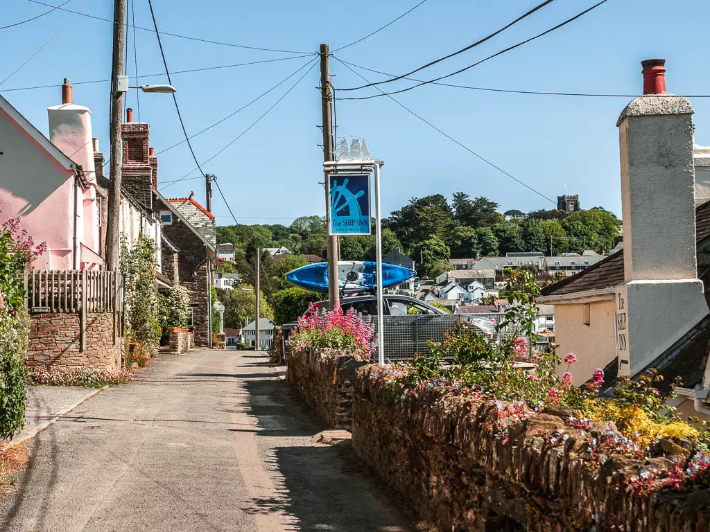 A road in Noss Mayo, with a brick wall on the right, with some pretty pink flowers. There are houses lining the road on the left, and the wall and roof of a pub in frame on the right.
