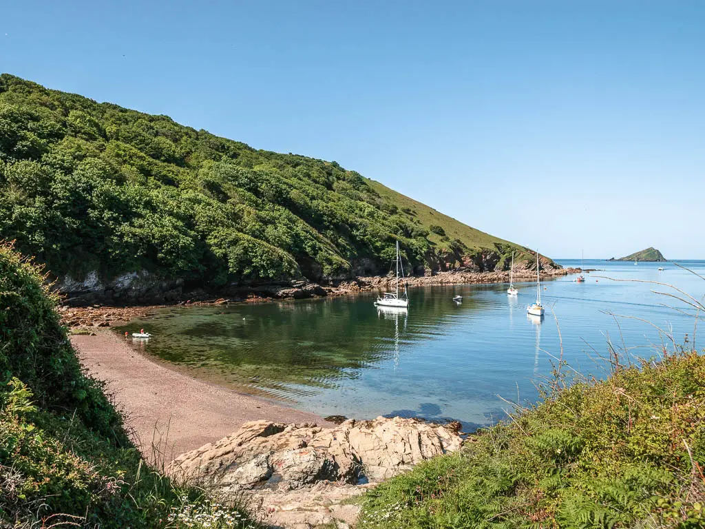 Looking down to a beach cove on the coastal walk from Noss Mayo.The beach is surrounded by tree a tree covered hill, and there are a few sail boats in the water.