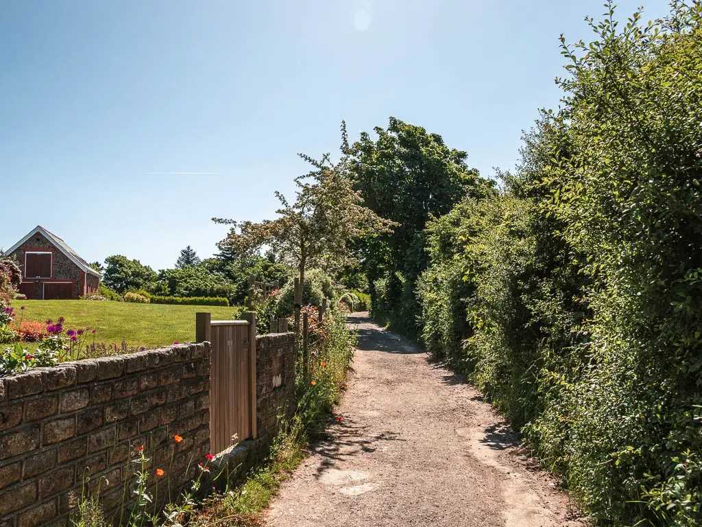 A road running straight, with hedges on the right and a stone and brick wall on the left. There is a person garden on the other side of the wall.