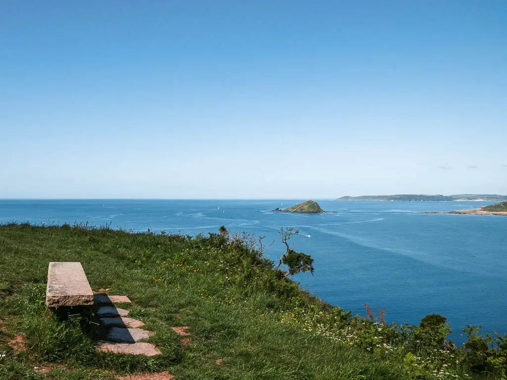 Looking across the grass hill top to the blue sea ahead and below. There is a small rock island in the sea, and a wooden bench on the hill to the left.