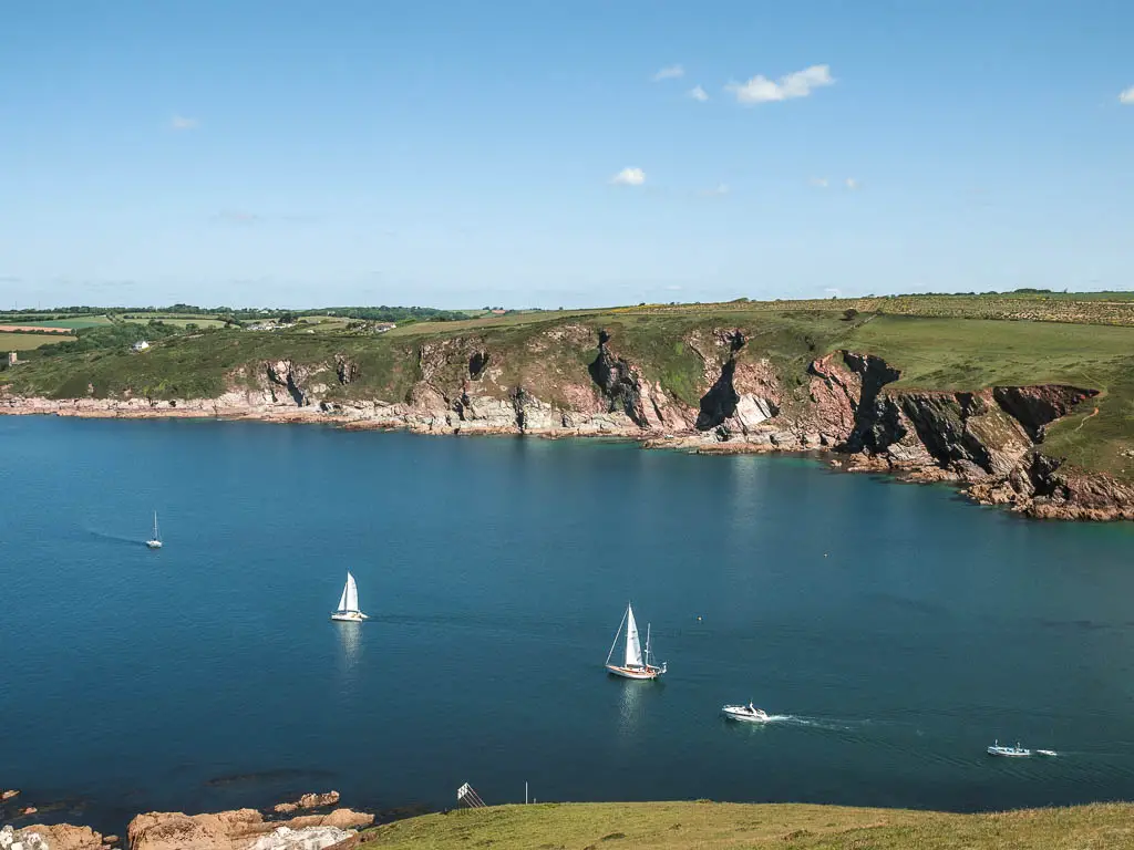Looking down to some small boats in there deep blue sea on the coastal Noss Mayo walk. There are rugged cliffs with a grass clifftop, on the other side of the sea.