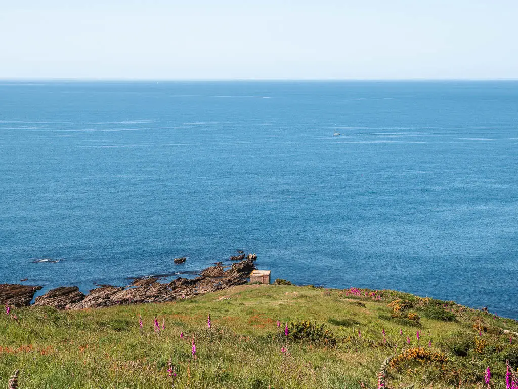 Looking down the grass hill to a small hut at the bottom on the coastal walk from Noss Mayo. Past the hut is the blue sea, as far as the eye can see. There are a few pink flowers in the grass.