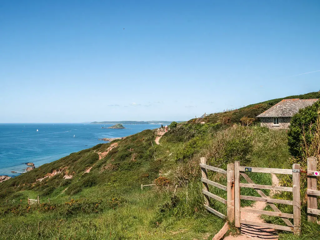 A wooden gate on the right, and a grass hill ahead and to the left, leading down to the blue sea.