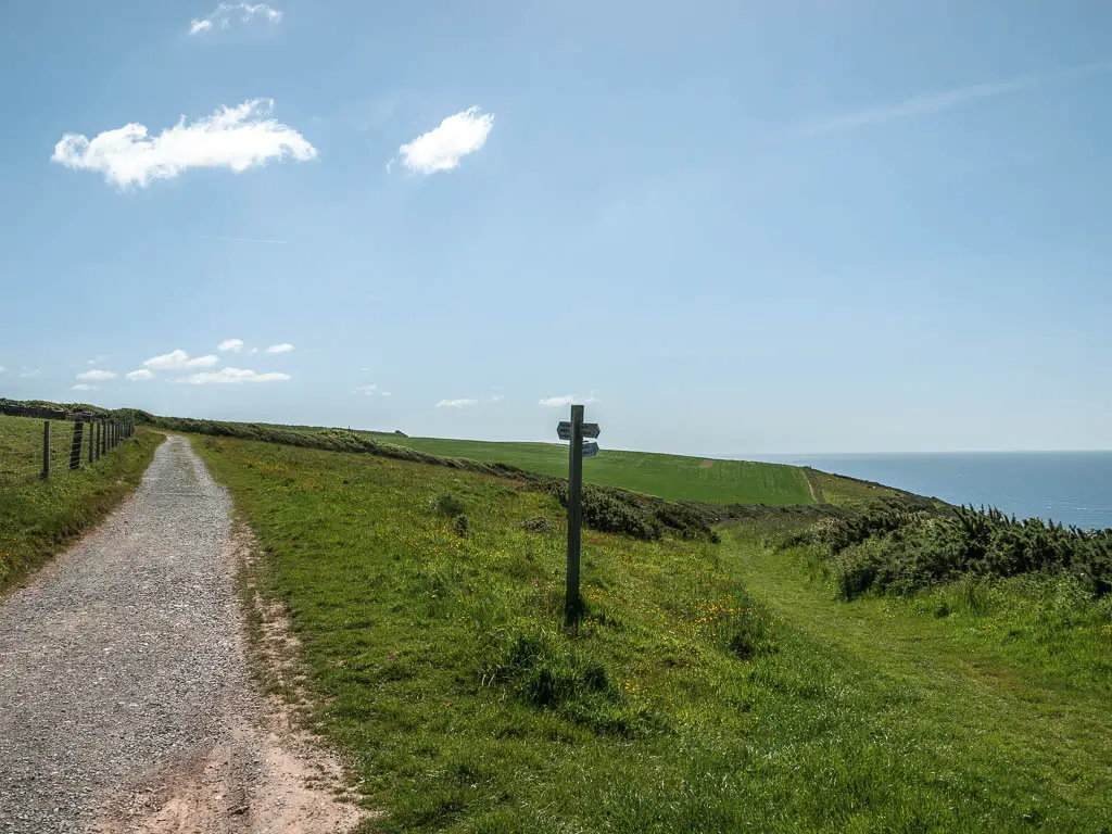 A gravel path on the left and a grass downhill, and grass path to the right. There is a wooden trail signpost between the path and the trail.