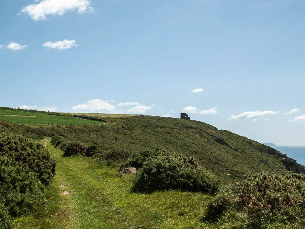 A grass trail on the left, running along the side of the hill. There are some ruins in the distance on top of the hill.