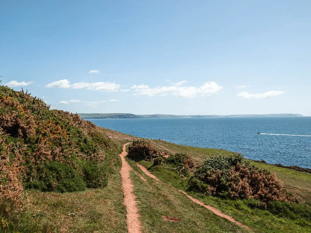A narrow trail along the side of a grass hill, with a few bushes dotted about, and the blue sea ahead.