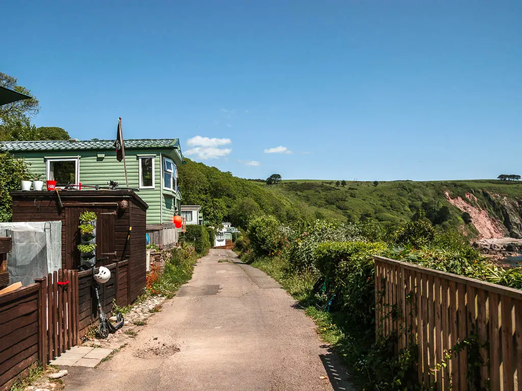 A path, with holiday trailer homes on the left and a wooden fence and bushes on the right.