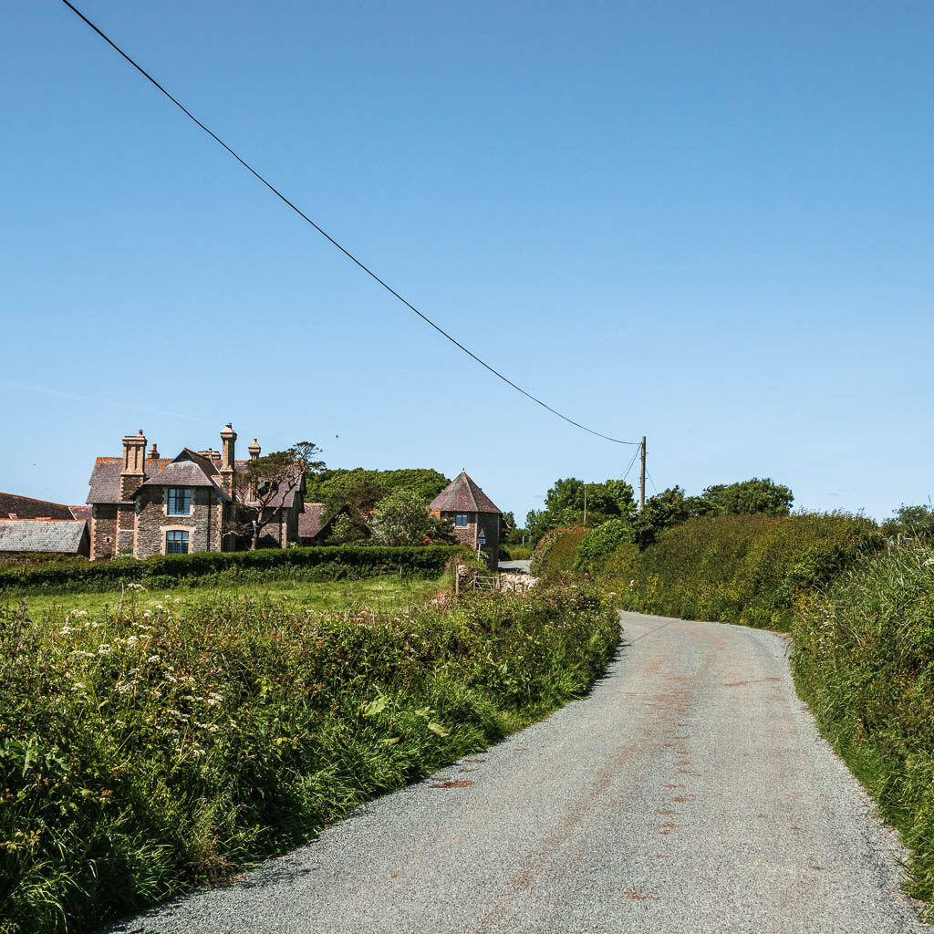 A country road, with a hedge on the right and smaller hedge on the left and large house ahead on the left.