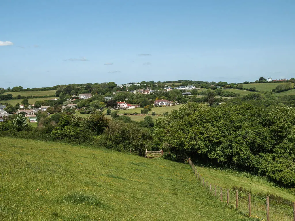 Looking down the grass hill, to a mass of trees at the bottom, and village hoses visible on a hill beyond that.