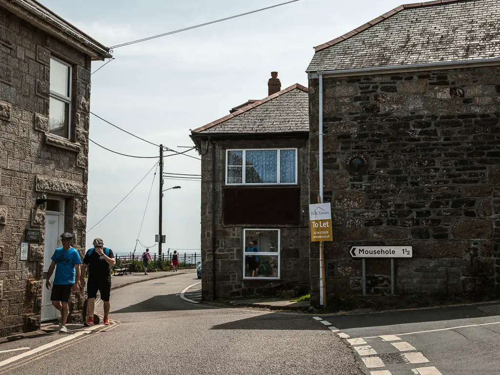 A road leading through a gap in the stone walled buildings. There is a road sign on the right, pointing left to Mousehole. There are a couple of men walking on the road. 