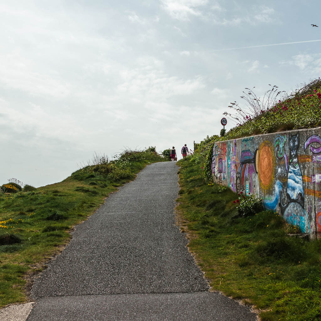 A path leading uphill on the walk from Penzance to Mousehole. The path is lined with grass, with a graffitis covered wall on the right. There are a couple of people walking at the top of the road. 